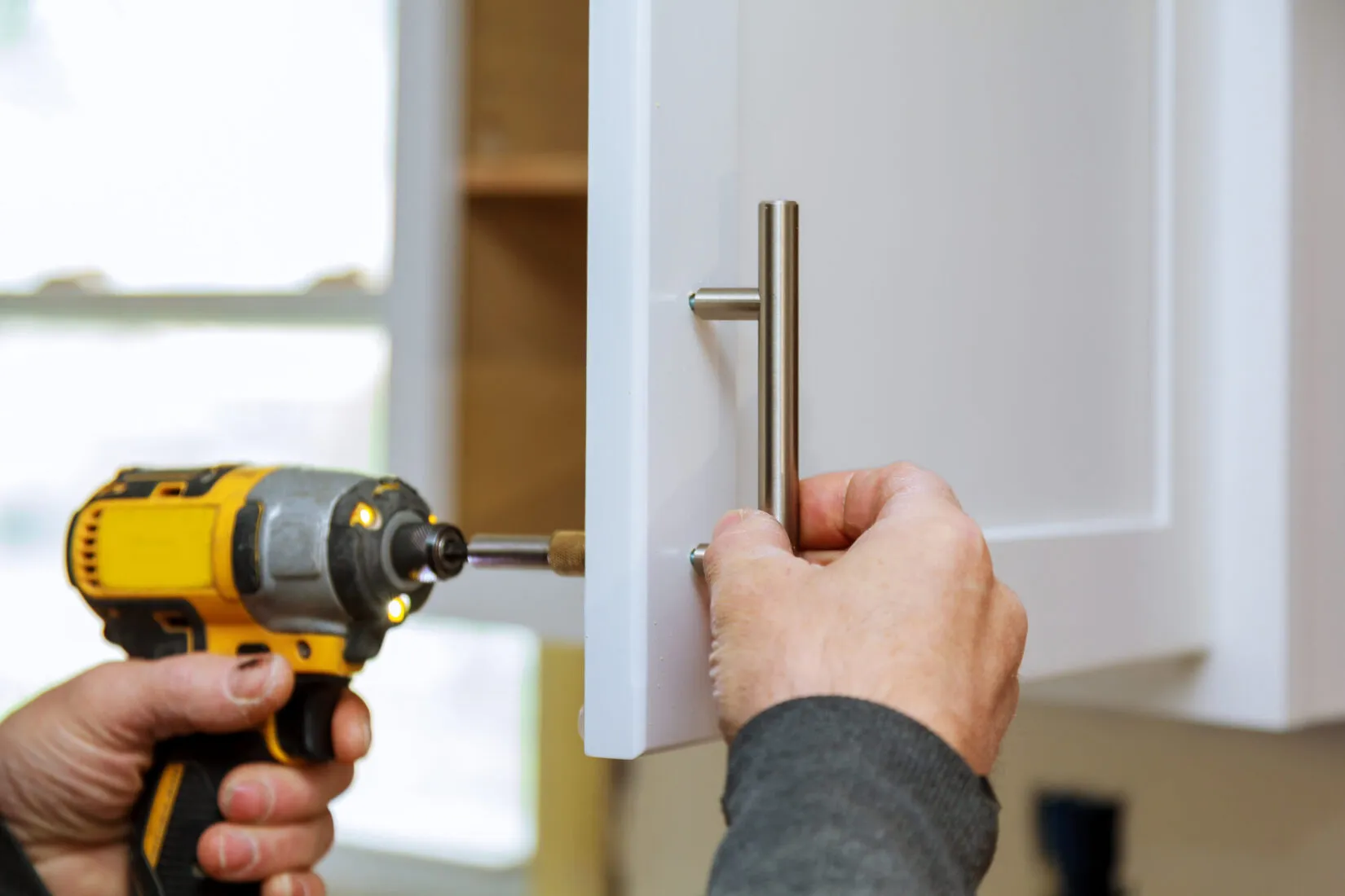 Person using a yellow cordless drill to install a silver handle on a white cabinet door.