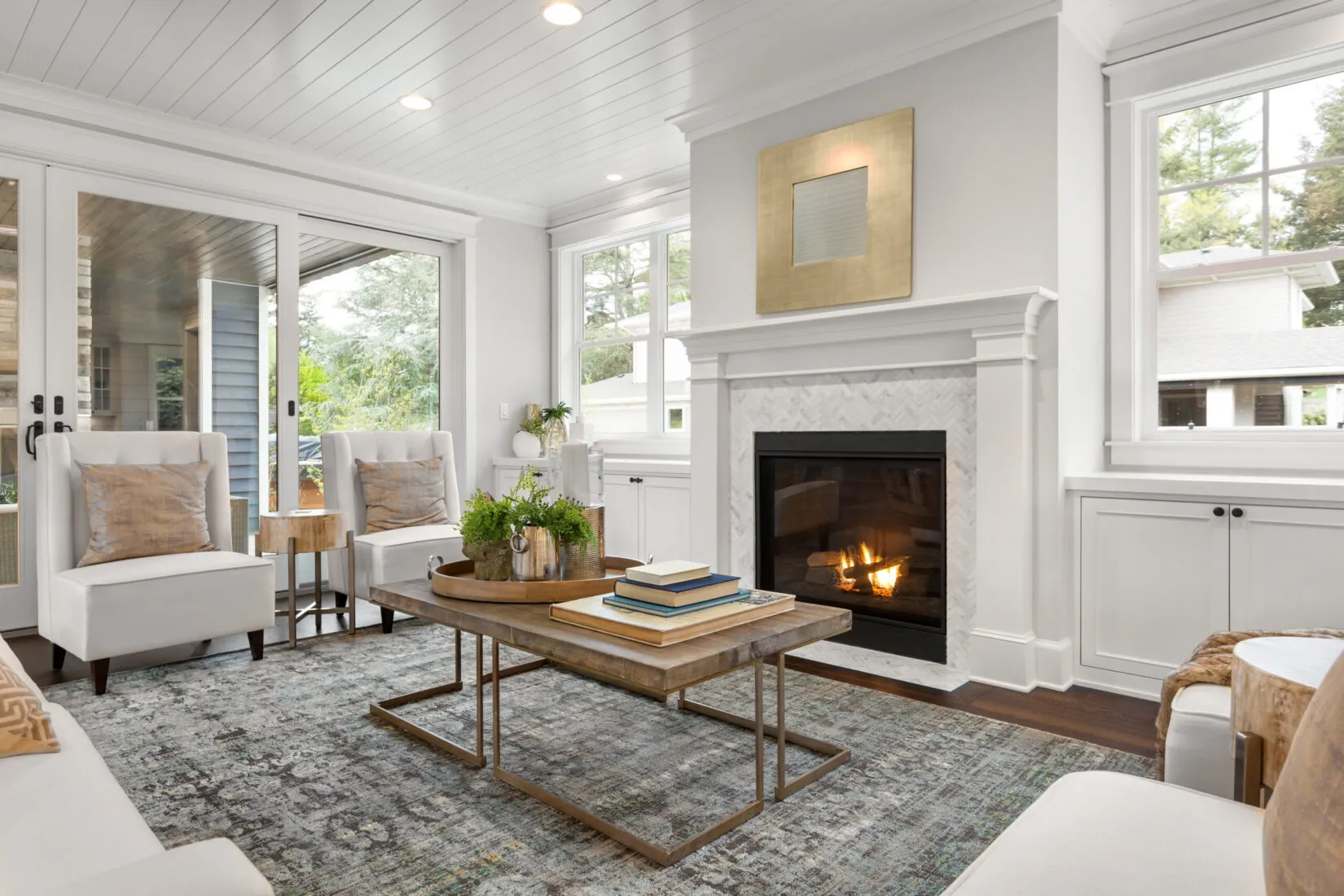 Bright living room with white armchairs, wooden coffee table with books and plants, and a lit fireplace under a gold-framed mirror.