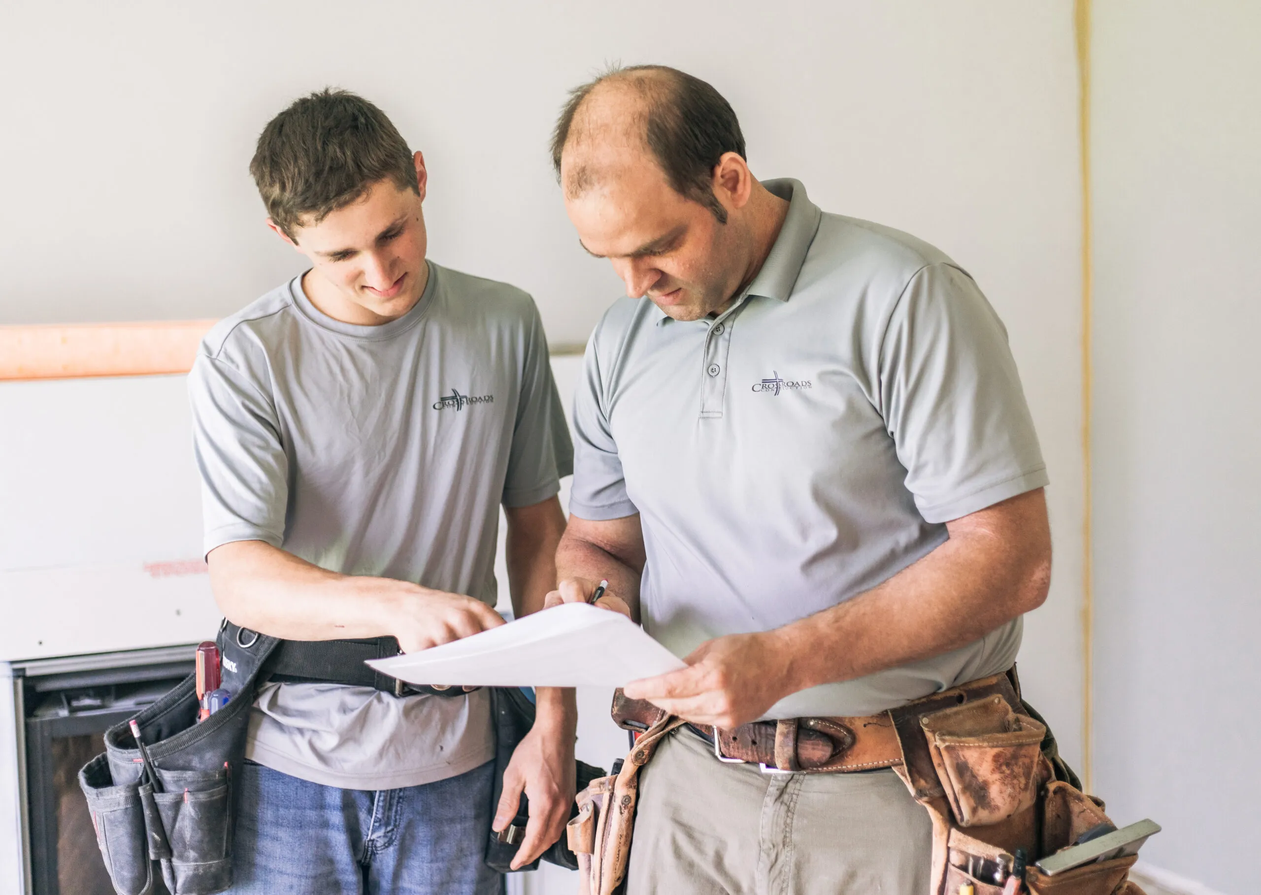 Two construction workers in gray shirts reviewing documents together indoors.