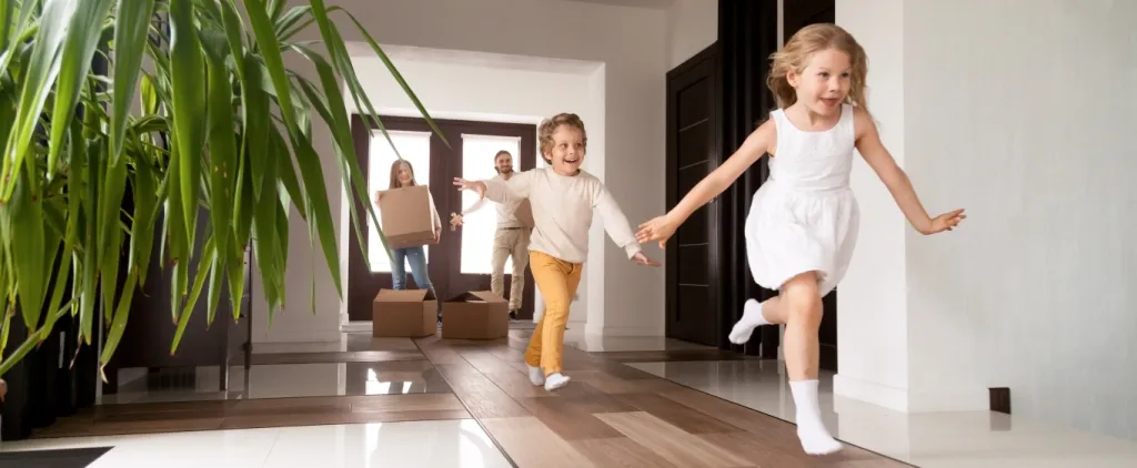 Two children running joyfully inside a new home while a man and woman carry moving boxes in the background.
