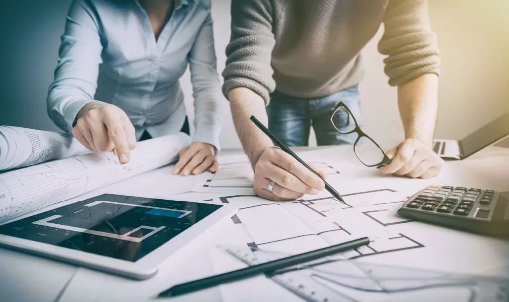 Two people working on architectural blueprints with a tablet, calculator, and drawing tools on the table.