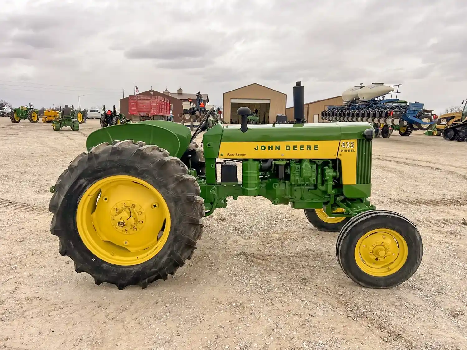 A John Deere tractor parked on a gravel lot with buildings and other machinery in the background