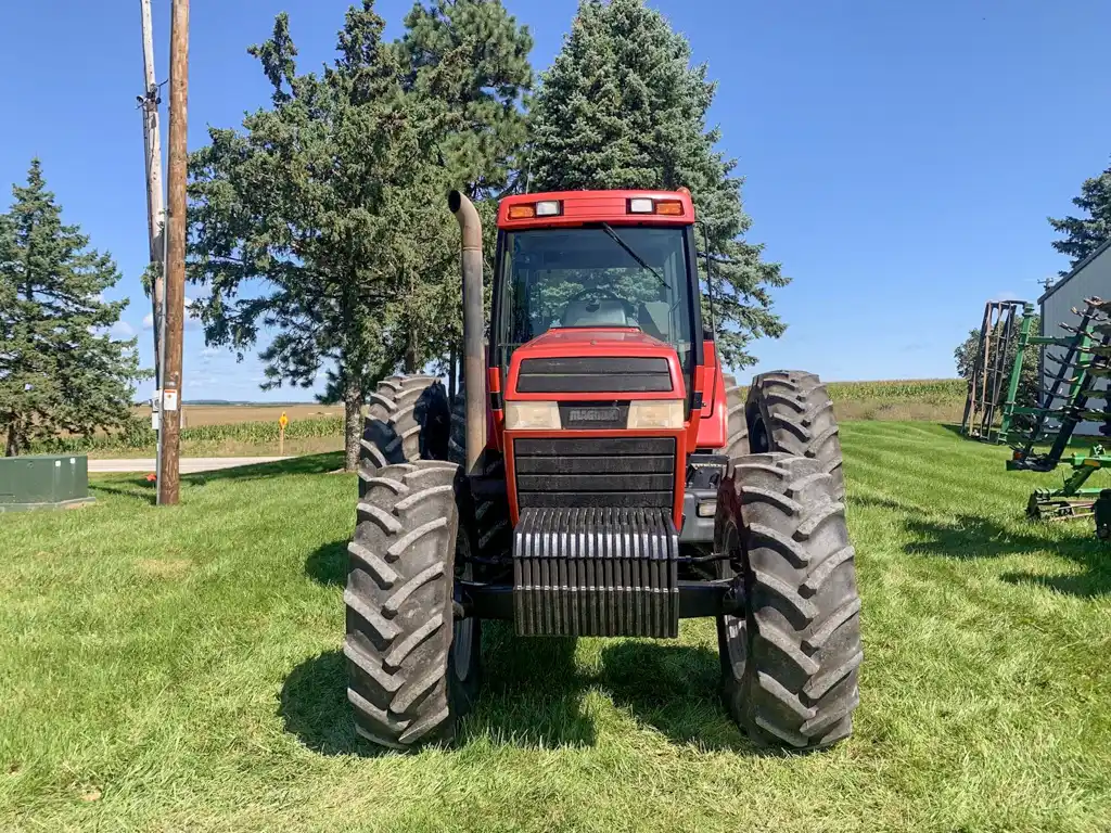 A red tractor facing directly forward parked on green grass with trees and a rural backdrop