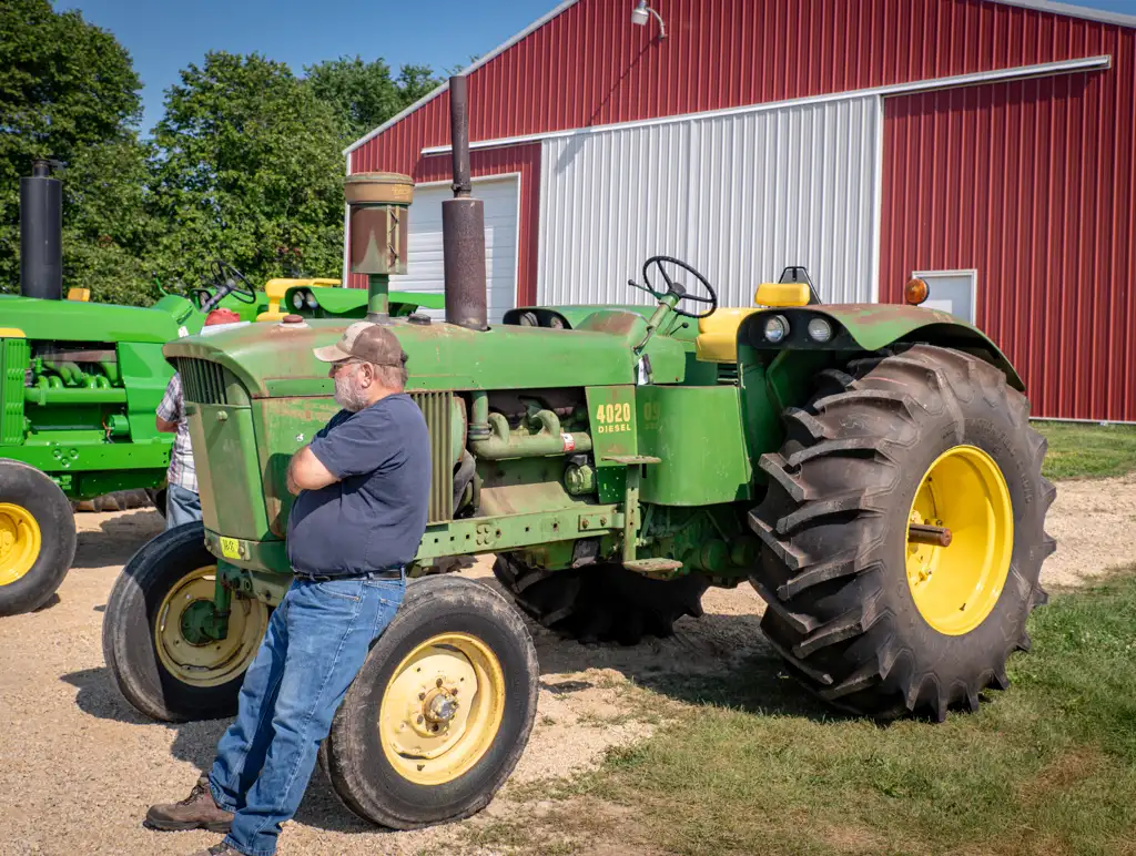 A man in a baseball hat leans against a John Deere 4020