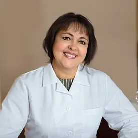 Smiling woman with dark hair wearing a white lab coat against a neutral background.