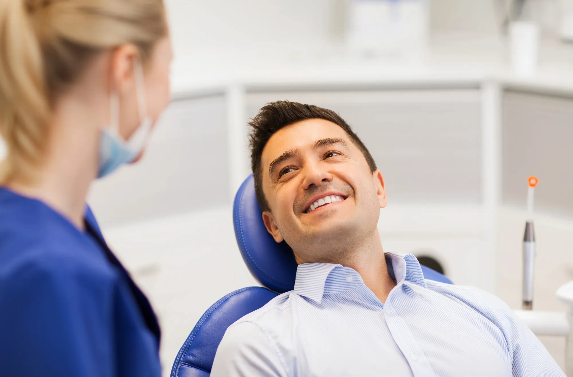 Smiling man reclining in a dental chair looking at a masked dental professional in blue scrubs.
