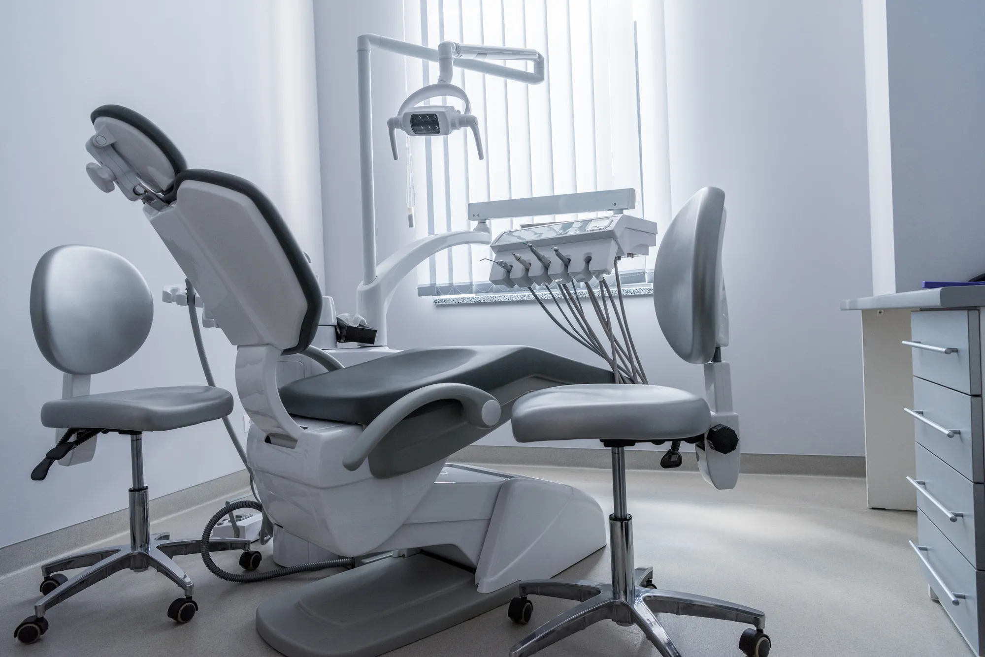 Modern dental exam chair with attached dental tools and two gray stools in a clean clinic room.