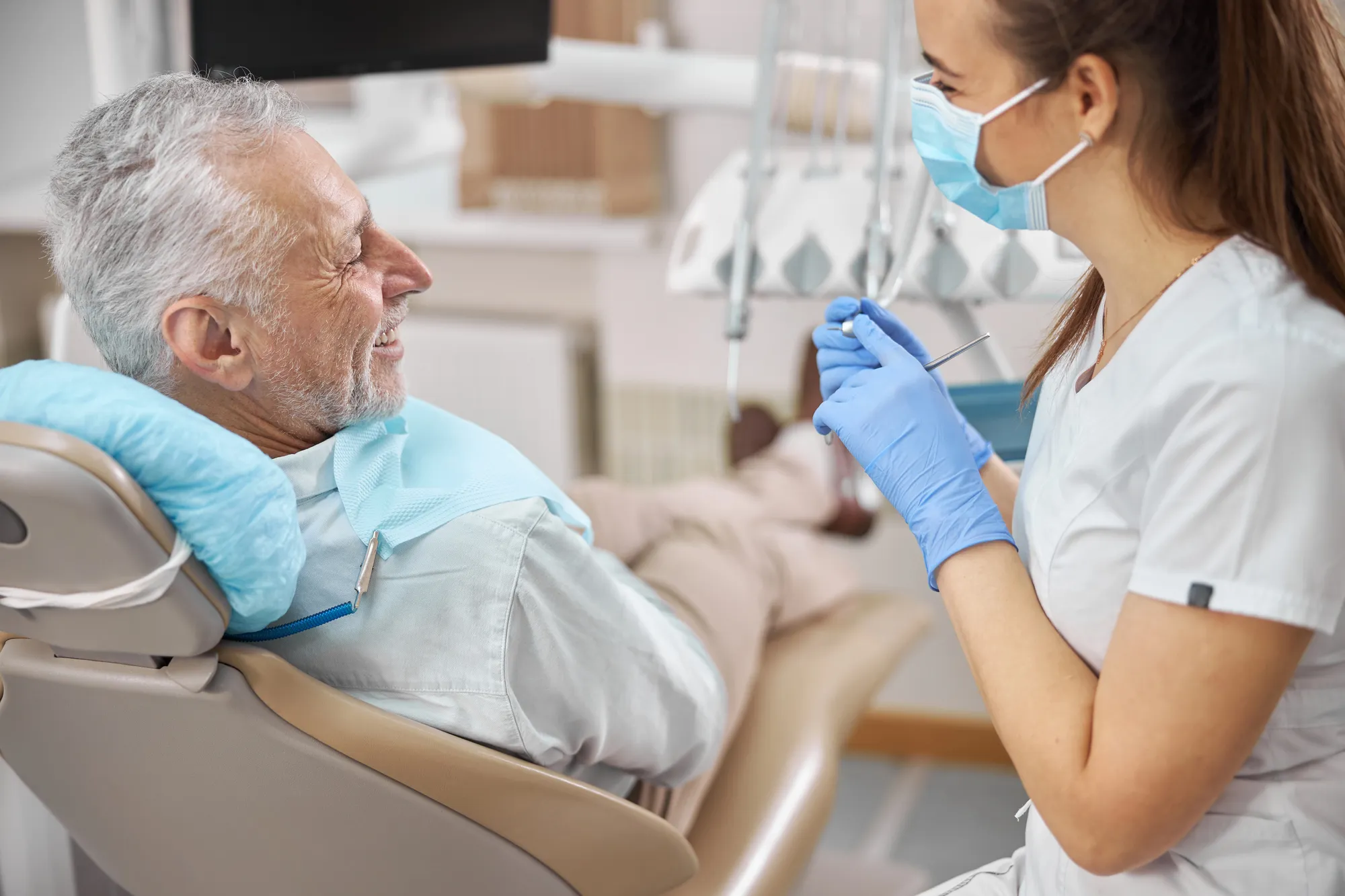 Dentist wearing mask and gloves smiles at a senior man reclining in a dental chair.