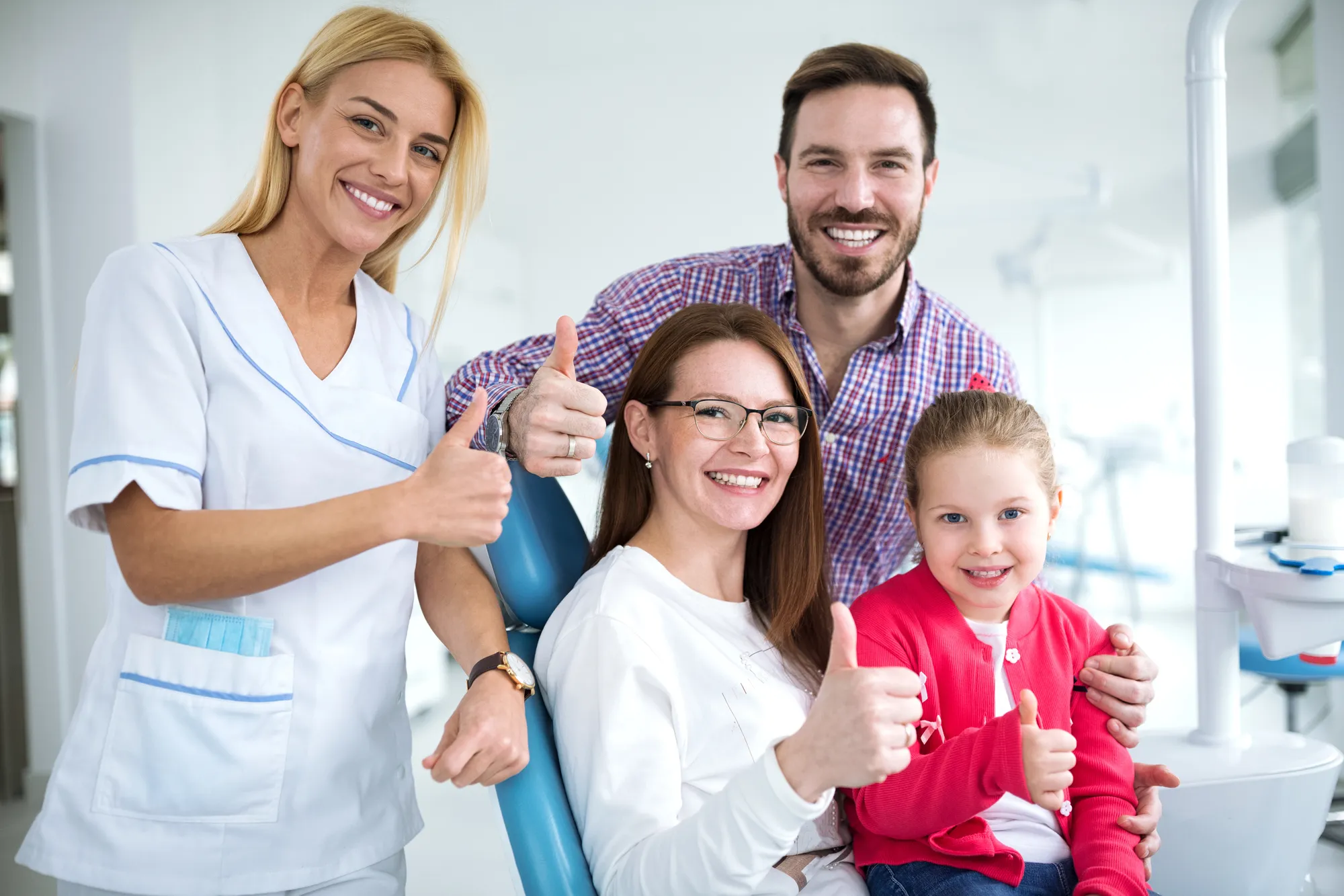 Smiling dentist, family, and child in dental office all showing thumbs up.