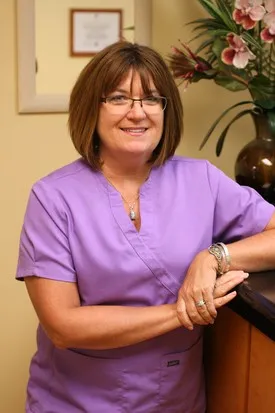 Smiling woman in purple scrubs with glasses posing indoors near a plant and framed diploma.