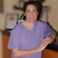 Smiling woman in purple medical scrubs with arms crossed in an indoor setting.