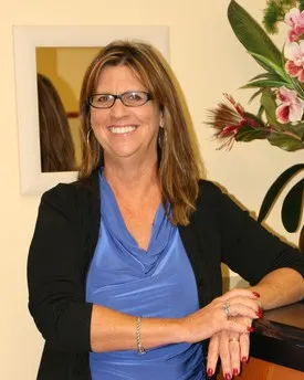 Smiling woman with glasses wearing a blue top and black cardigan, standing by a counter with flowers nearby.