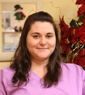 Smiling woman with long brown hair wearing a lavender scrub top indoors with red flowers behind her.
