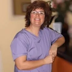 Smiling woman with glasses wearing a purple medical scrub top leaning on a counter indoors.