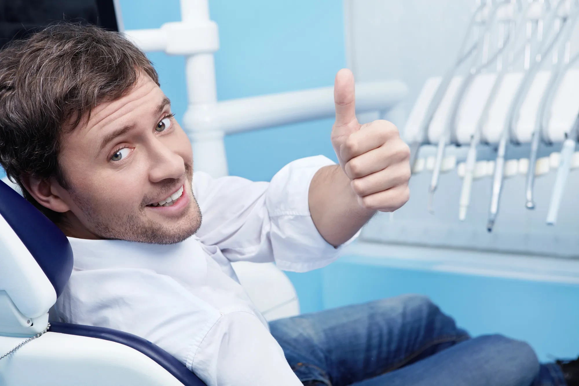 Smiling man sitting in dental chair giving thumbs up with dental tools in background.
