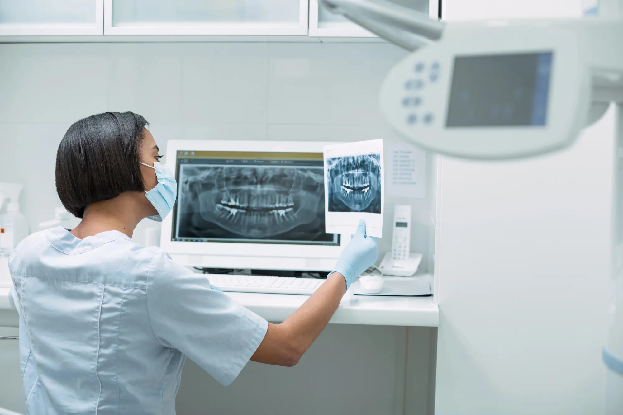 Dental professional wearing a mask and gloves examines a dental X-ray image by a computer screen.