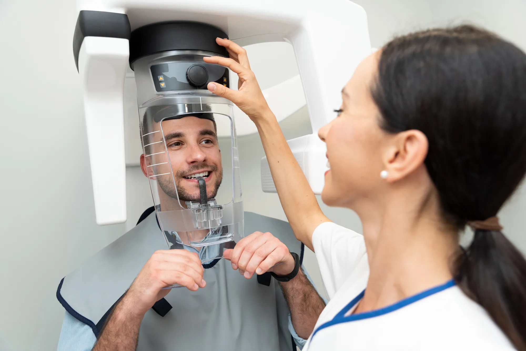Technician adjusts a dental X-ray machine while a man bites a plastic dental guide.