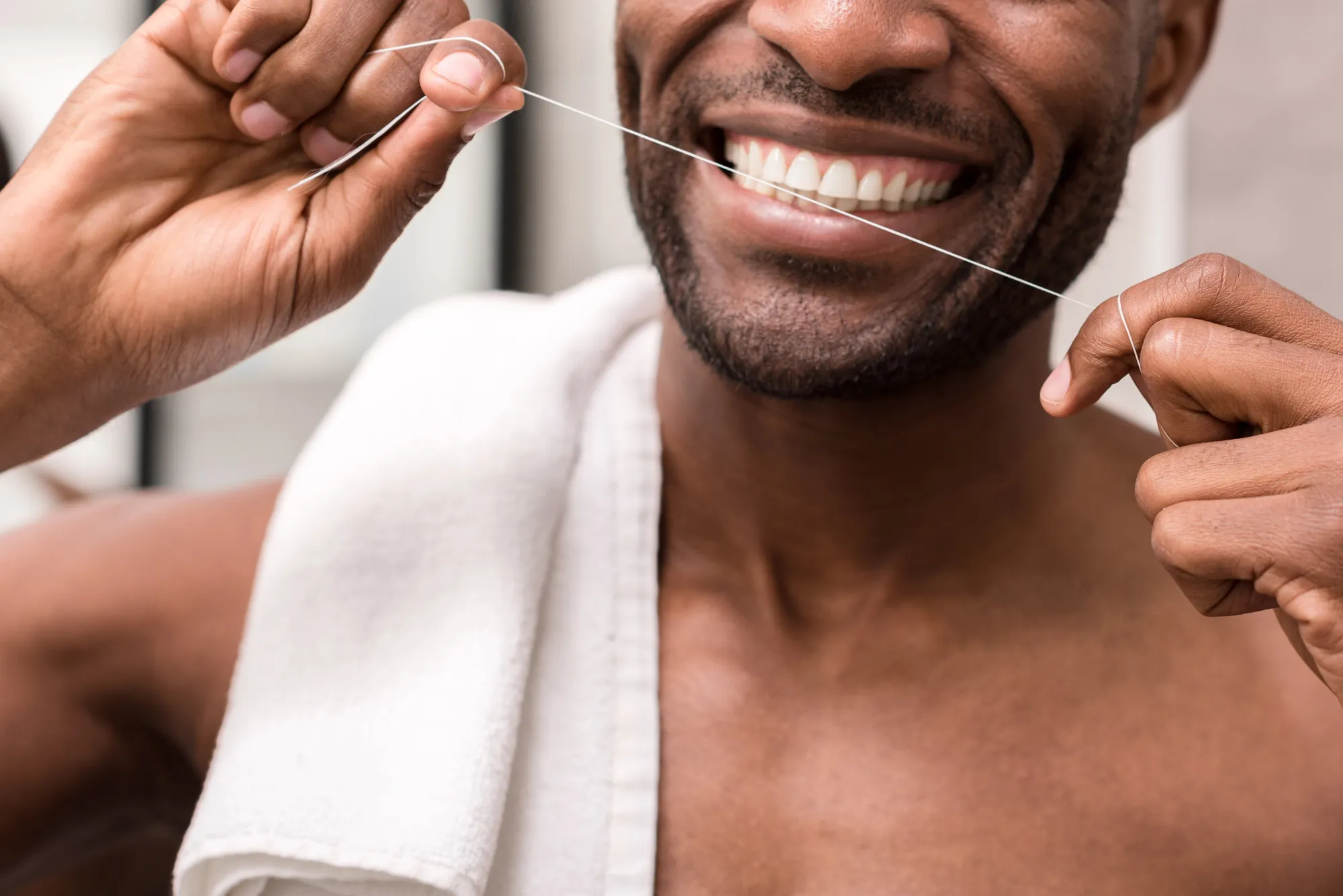 Smiling man with a white towel on his shoulder flossing his teeth with dental floss.