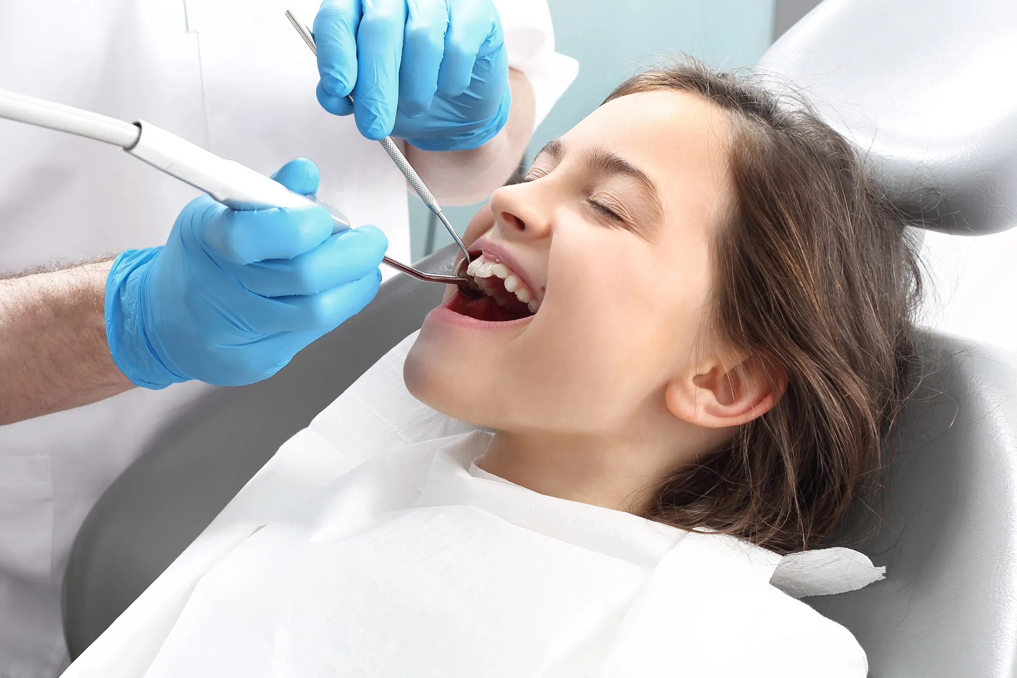 Dentist wearing blue gloves examining a young girl's teeth with dental tools during checkup.