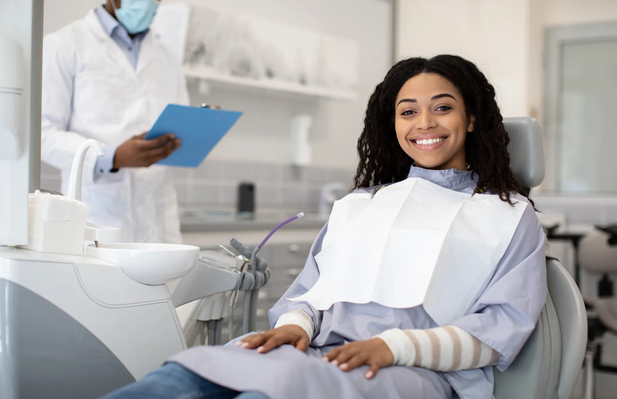 Smiling woman wearing dental bib sitting in dentist chair with dentist holding clipboard in background.