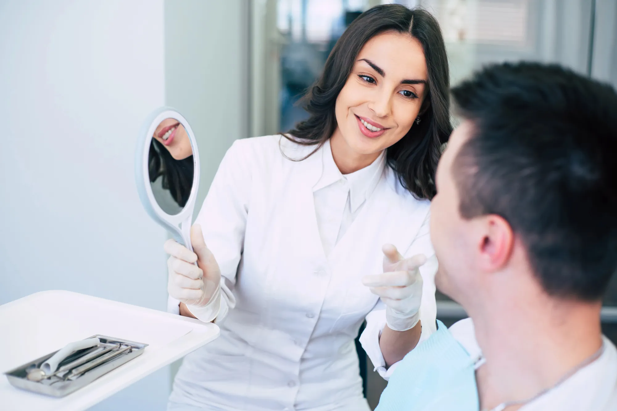 Female dentist in white coat holding a mirror and smiling at a male patient in dental clinic.