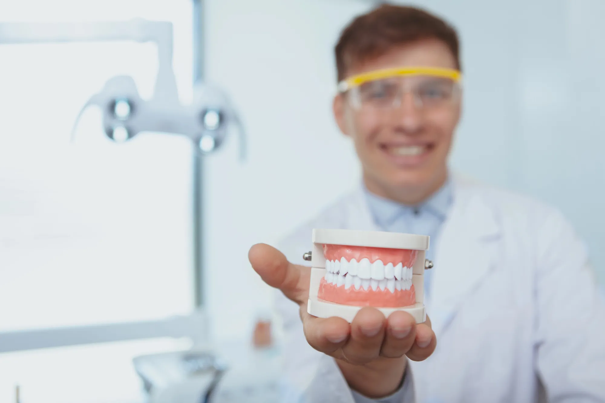 Dentist in white coat holding a dental model of teeth and gums toward the camera.