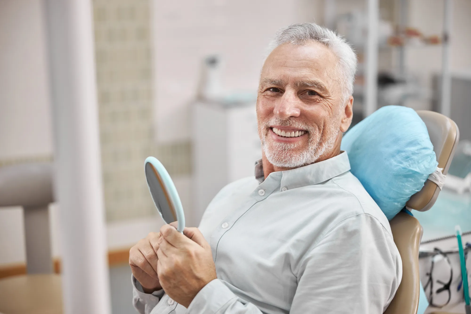 Smiling elderly man sitting in a dental chair holding a hand mirror in a dental clinic.