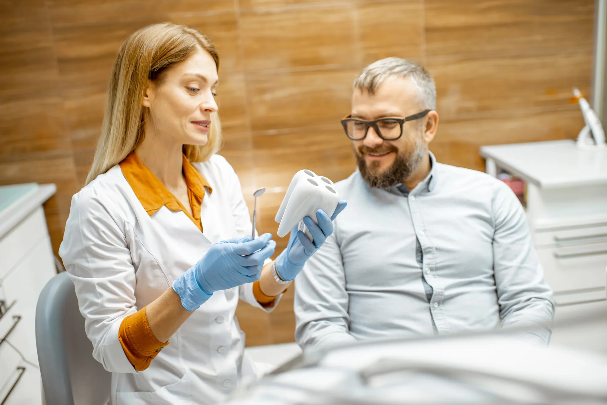Dentist in gloves shows a dental tool and a tooth model to a smiling male patient in a clinic.