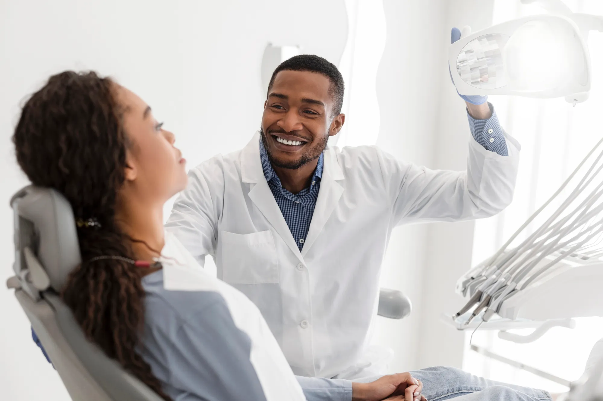 Smiling dentist adjusting light above patient seated in dental chair in bright clinic.