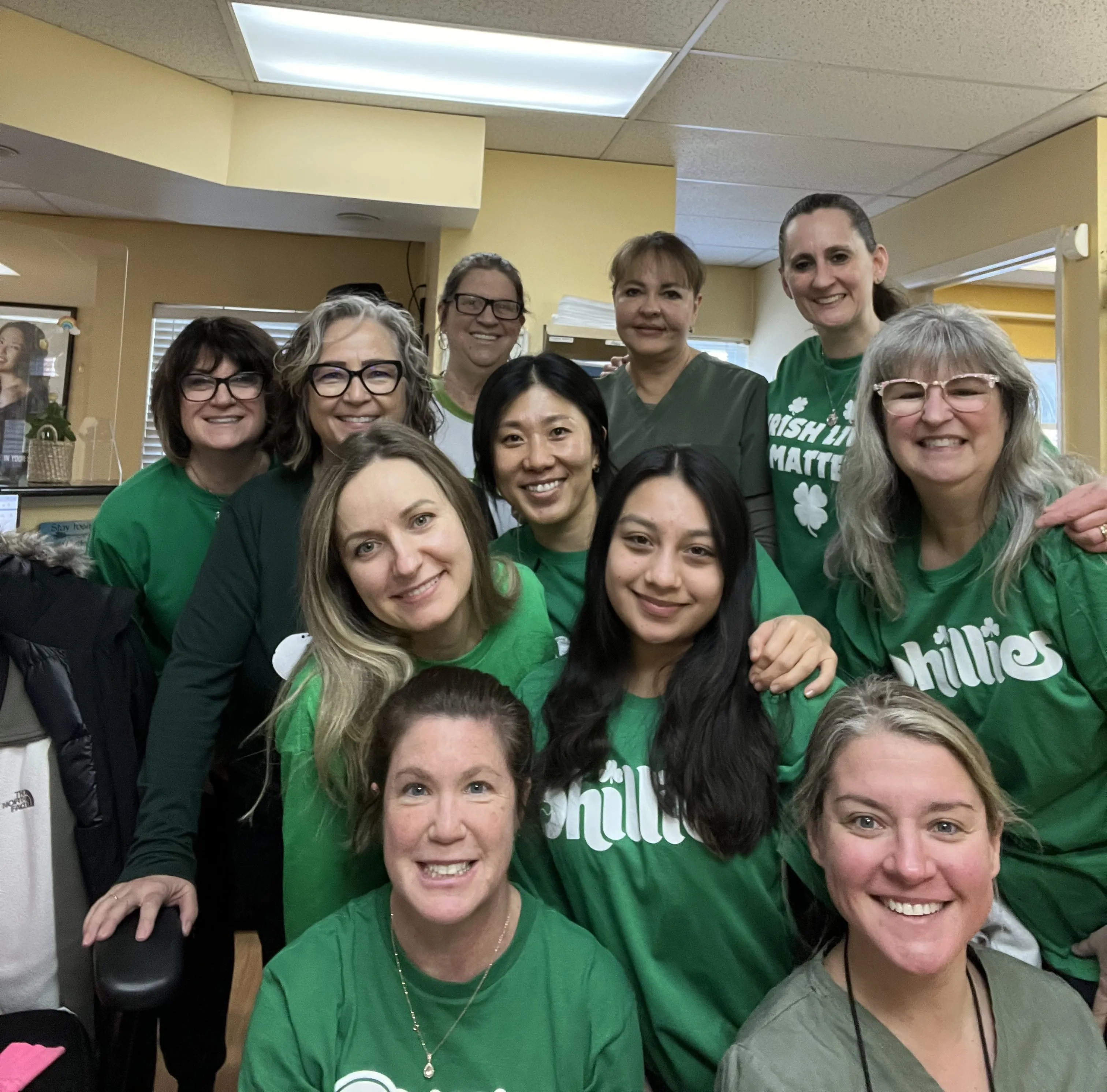 Eleven women smiling indoors, most wearing green shirts with text like Phillies and Irish in Matters.