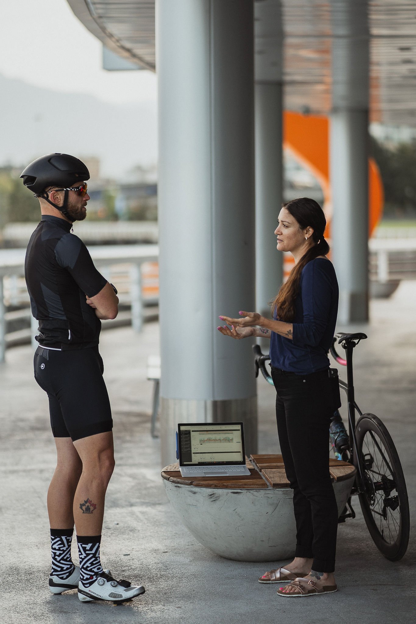 A cyclist in black gear and helmet standing and talking to a woman in casual clothes beside a bicycle and a laptop on a concrete bench.