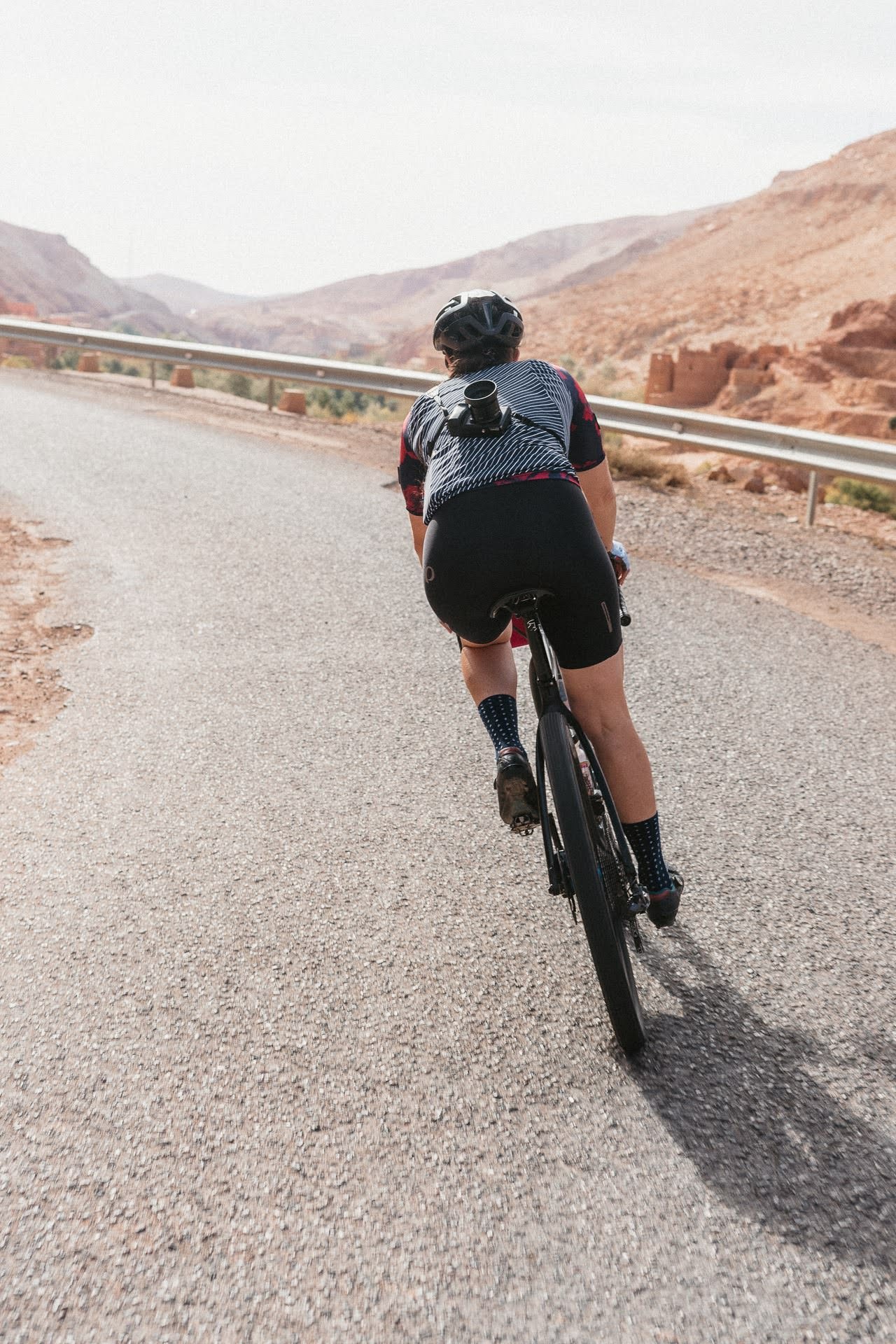 Cyclist wearing a helmet and black cycling shorts rides on a mountain road with a camera mounted on their back.