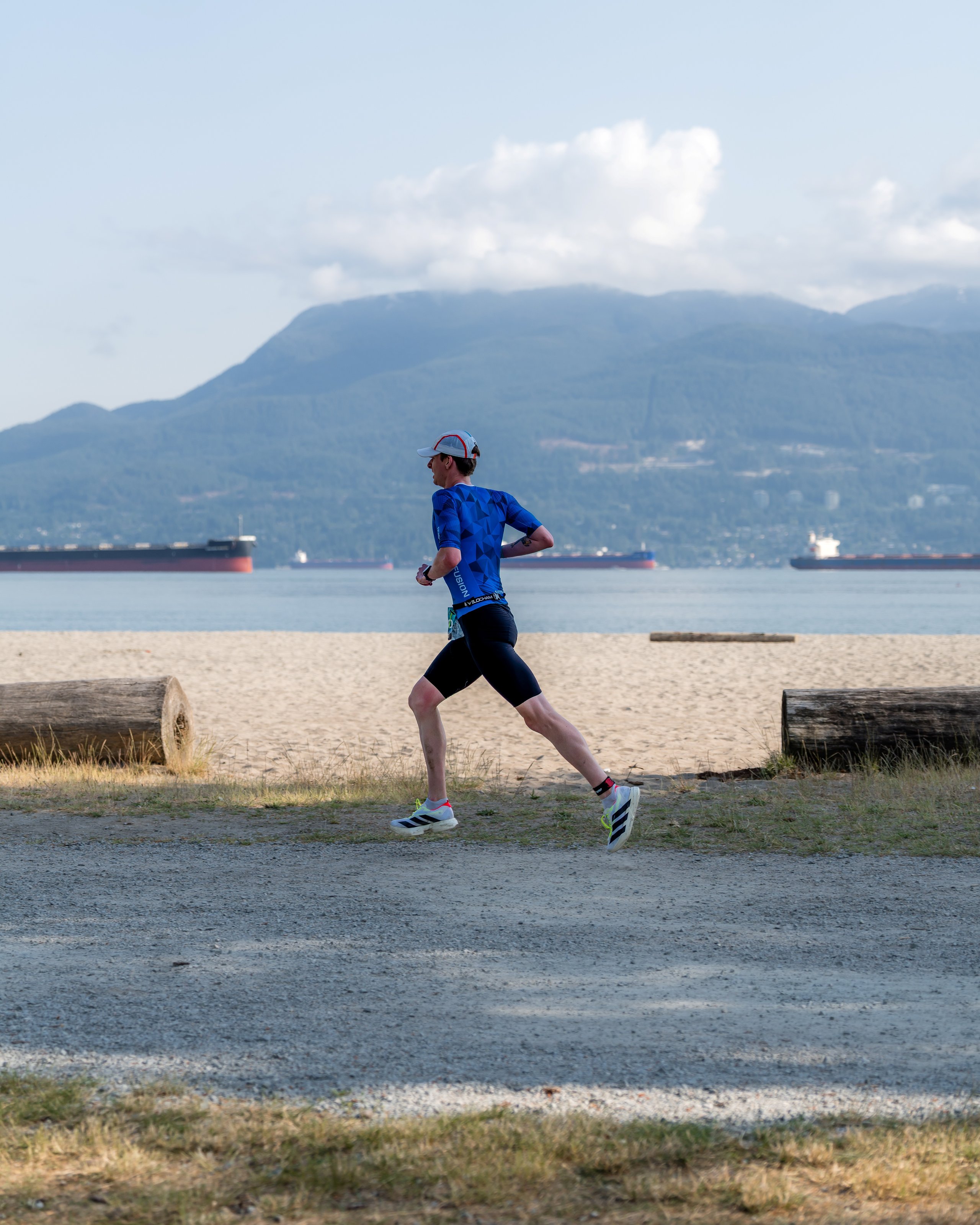 Runner dressed in blue athletic wear and cap running along a path by the beach with mountains and cargo ships in the background.