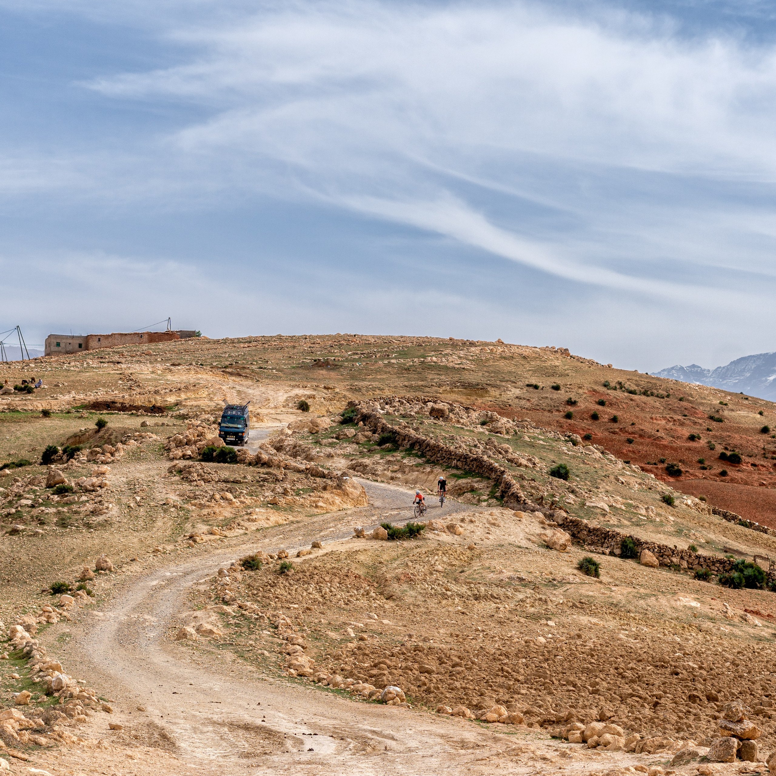 Two cyclists riding on a winding dirt road through a rocky hillside with a blue truck and stone walls nearby under a partly cloudy sky.