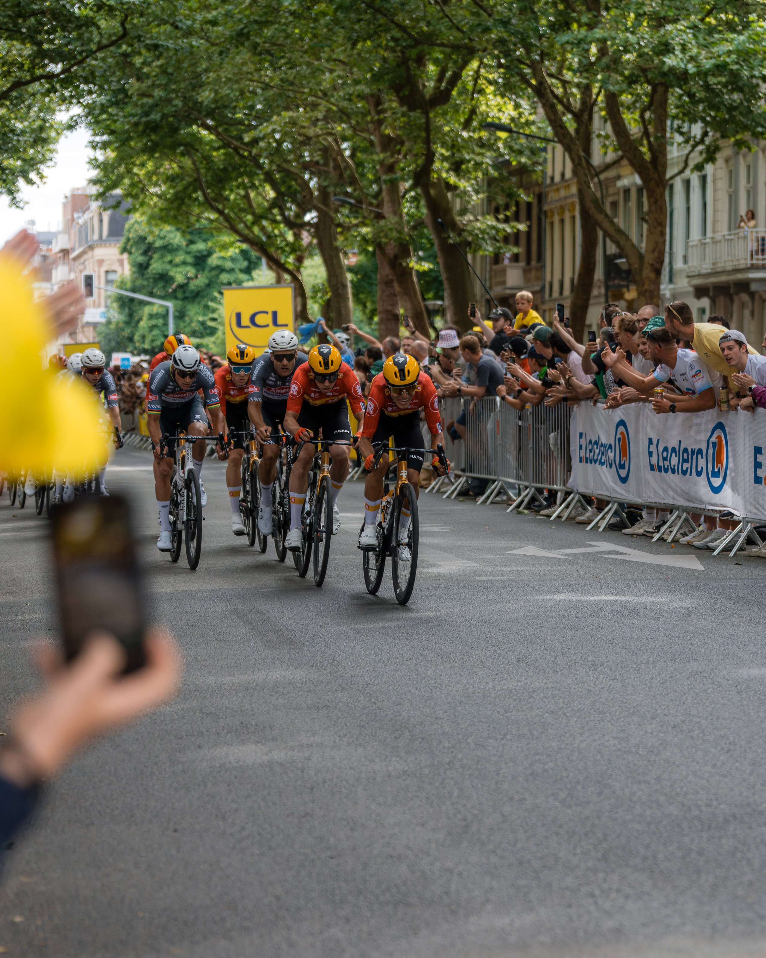 Cyclists in a road race riding closely past cheering spectators along a tree-lined street.