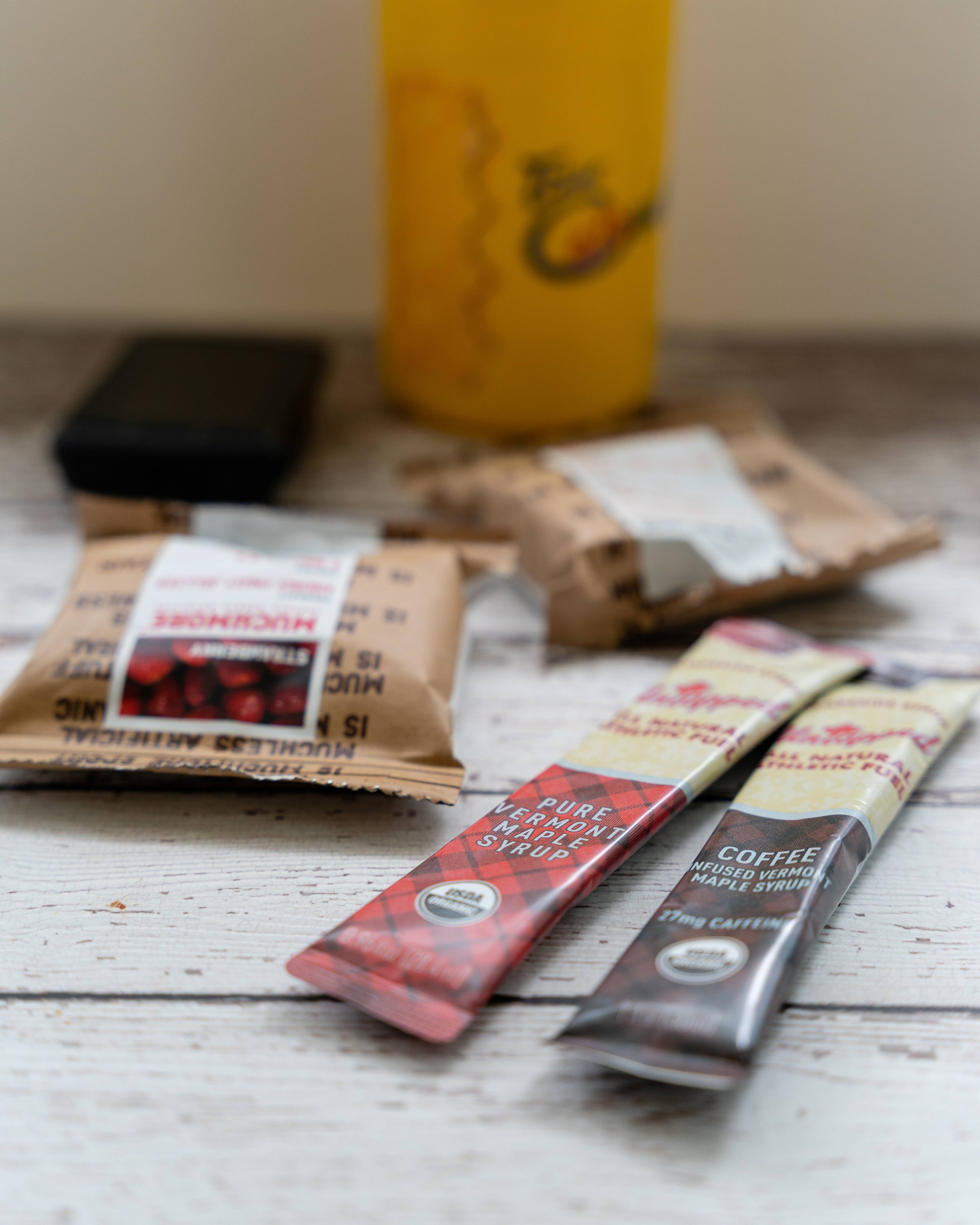 Two small packets of Vermont maple syrup, one pure and one coffee-infused, lying on a rustic wooden table with blurred packaging and a drink in the background.