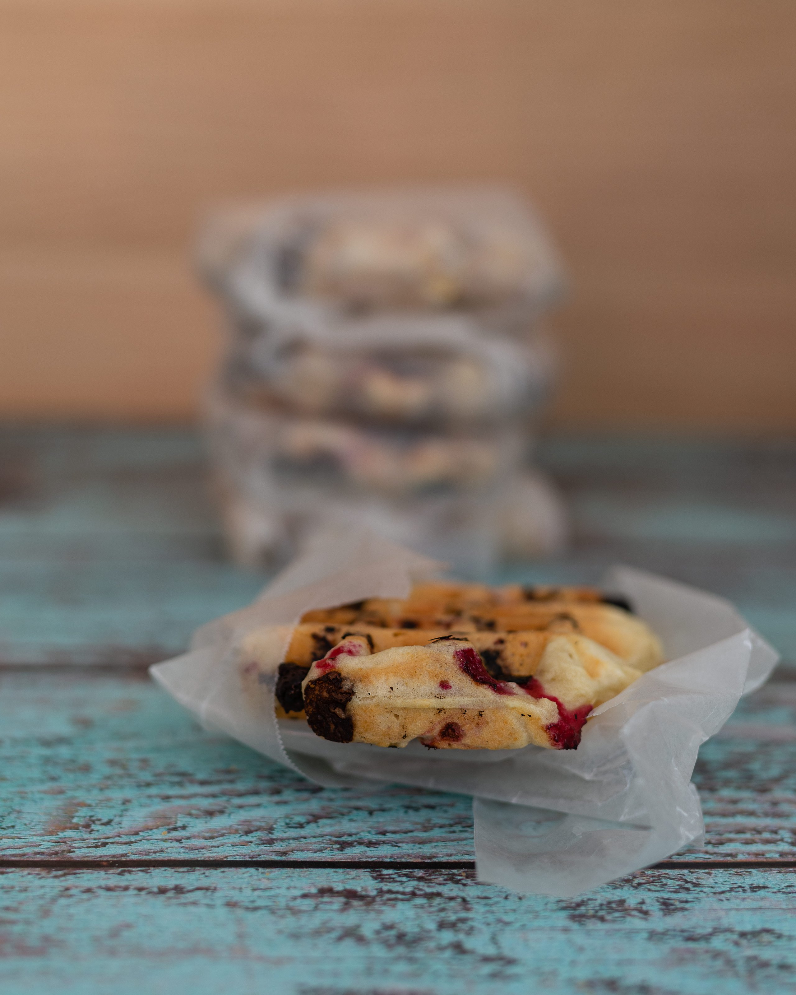 Close-up of a wrapped raspberry and chocolate chip cookie on a rustic blue wooden surface, with blurred stacks of cookies in the background.