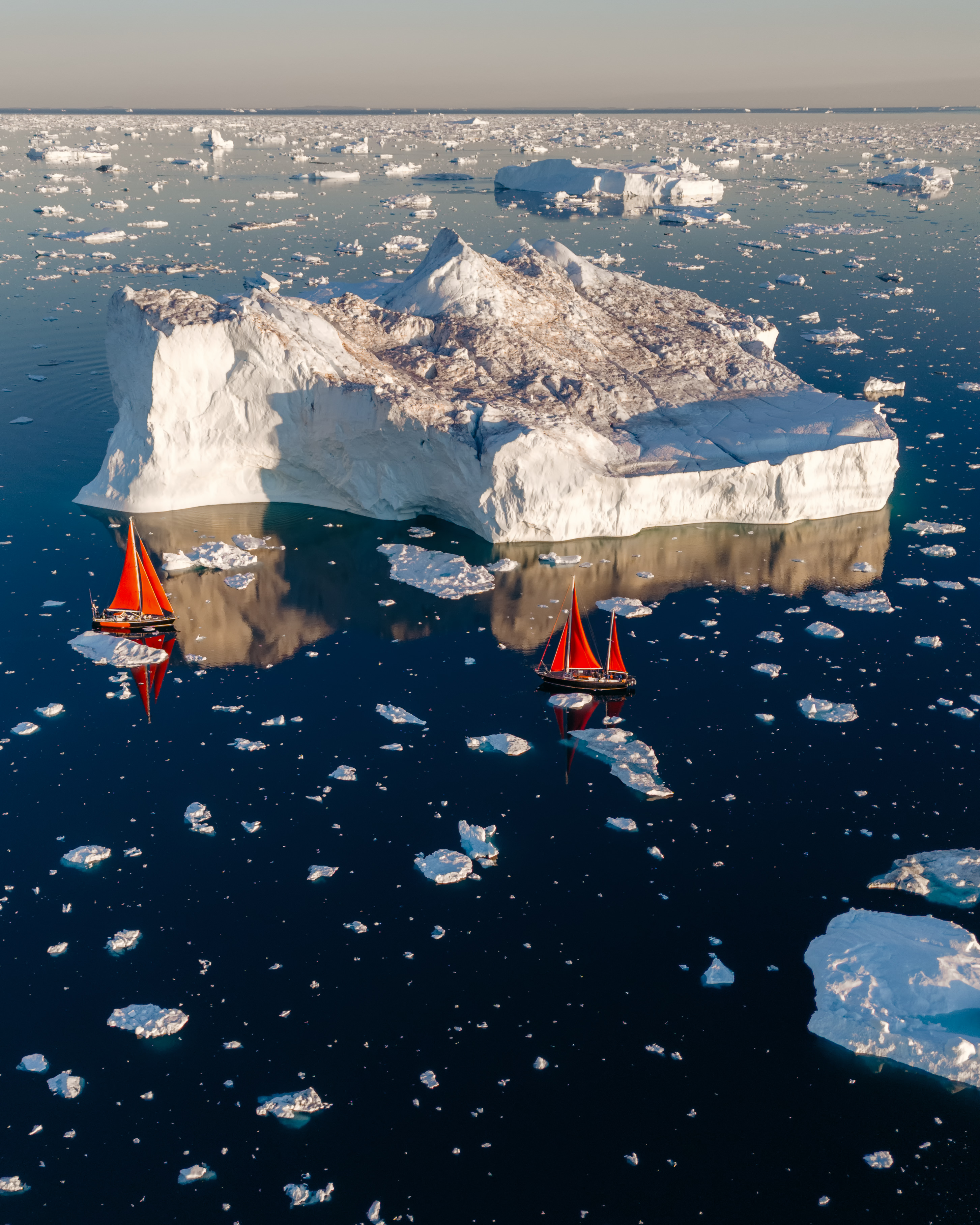 Two sailboats with red sails navigating icy waters near a large iceberg at sunset.
