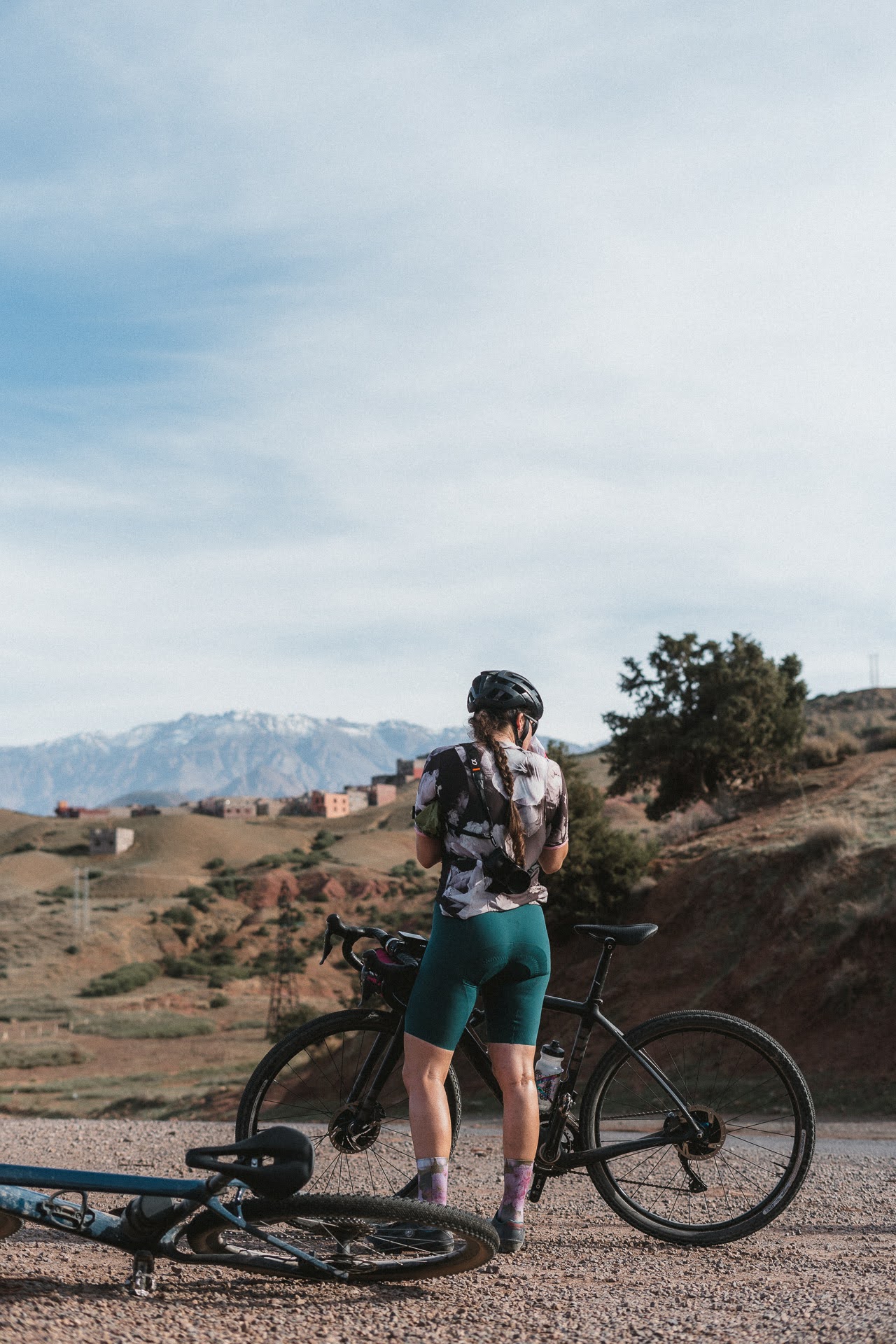 Cyclist in helmet and floral shirt standing with a black road bike on a gravel path with mountains in the background.