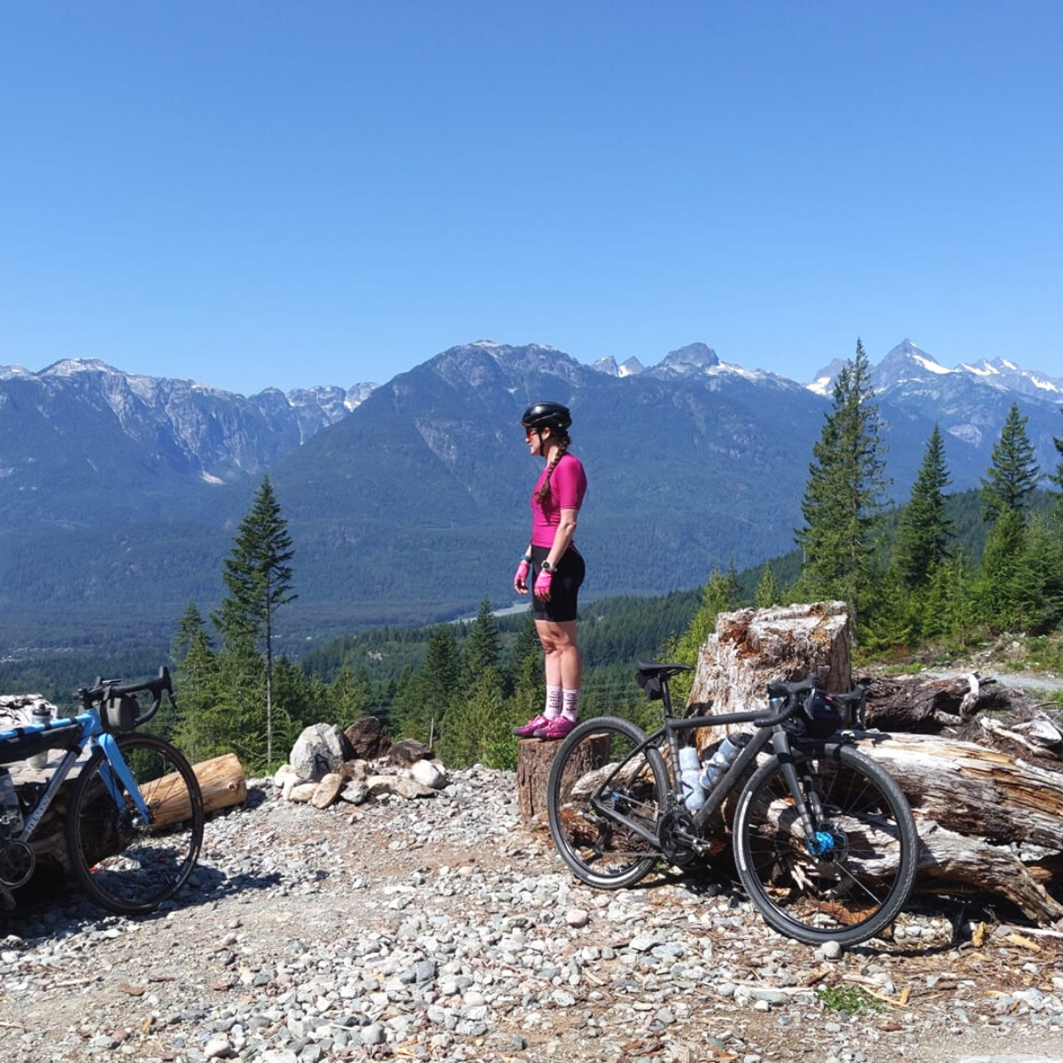 Woman in pink cycling outfit and helmet standing on a tree stump next to two parked bicycles with mountain and forest background under a clear blue sky.