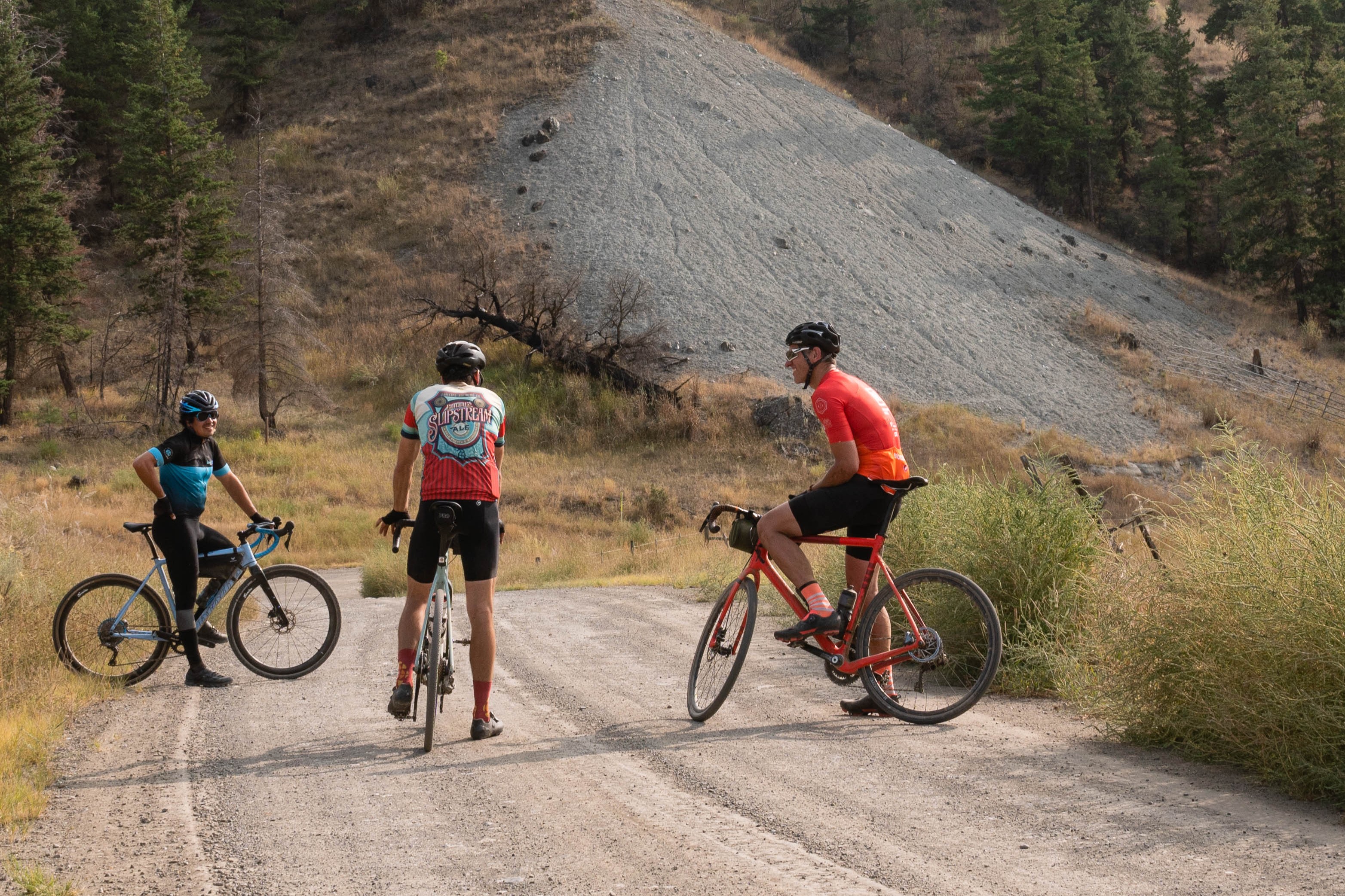 Three cyclists on gravel road in mountainous area with dry grass and trees, two sitting on bikes and one standing.