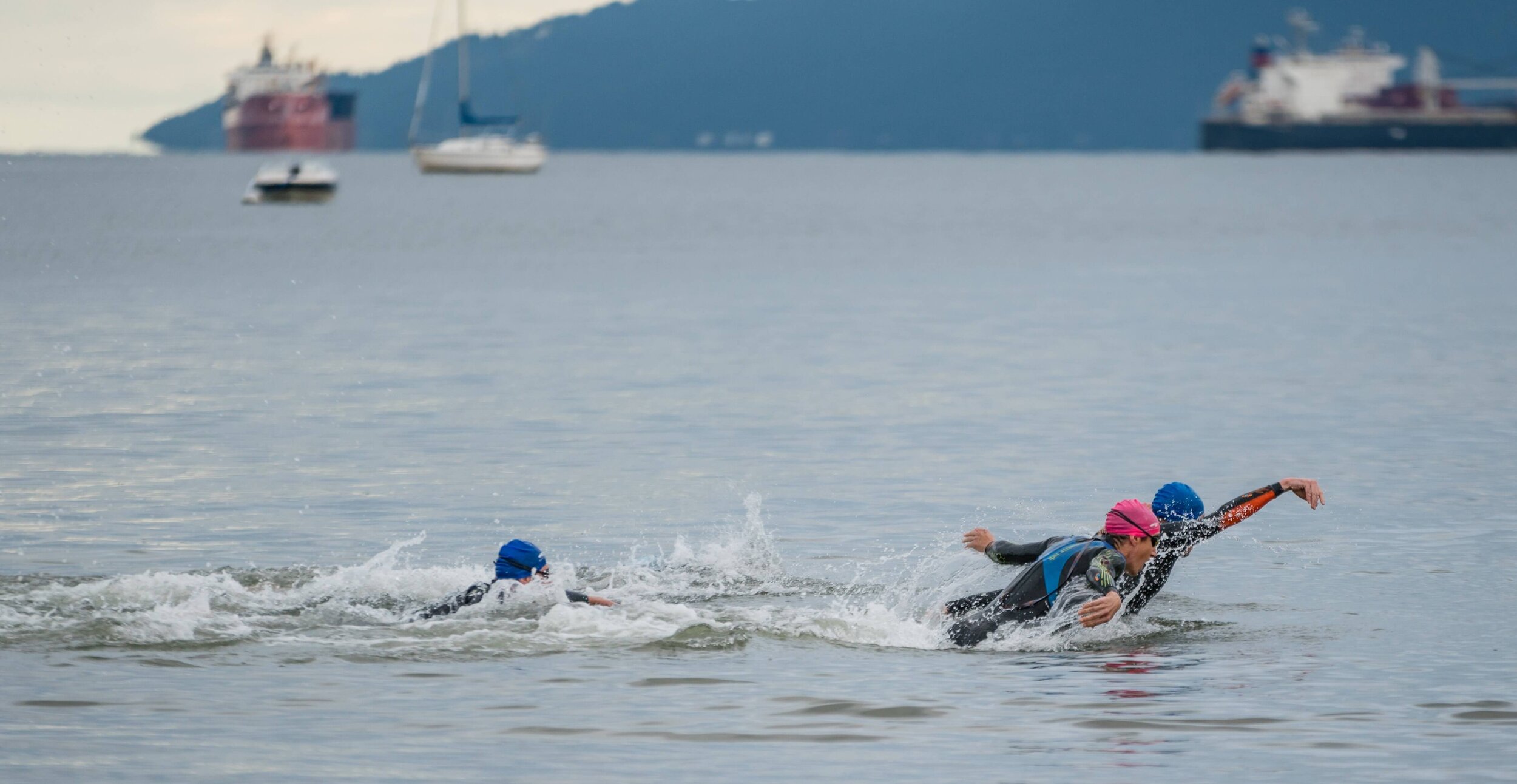 Three swimmers wearing wetsuits and swim caps racing in open water with boats and a mountain in the background.