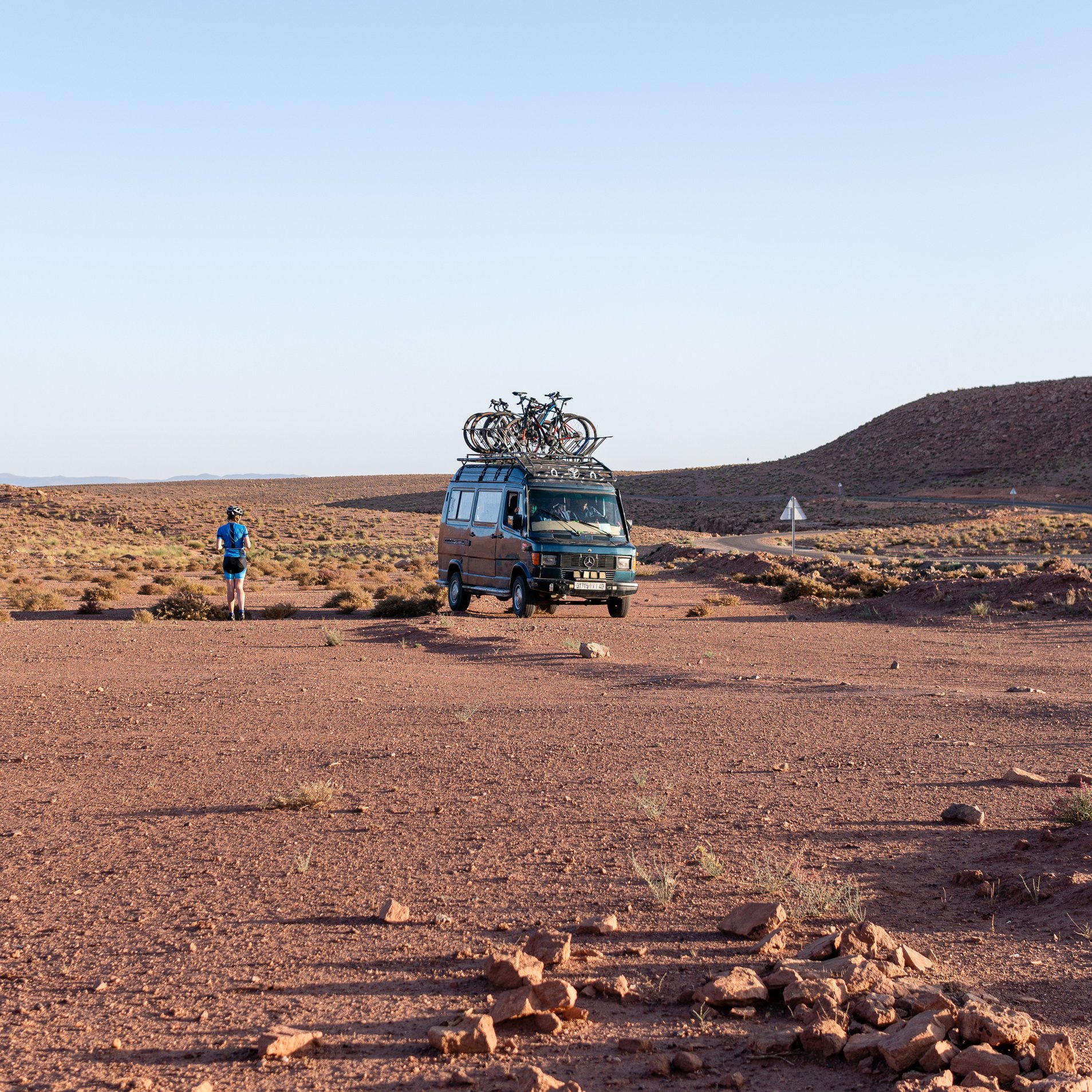 Black van with multiple bicycles mounted on its roof, parked on a vast, arid dirt plain with a cyclist standing nearby facing away.