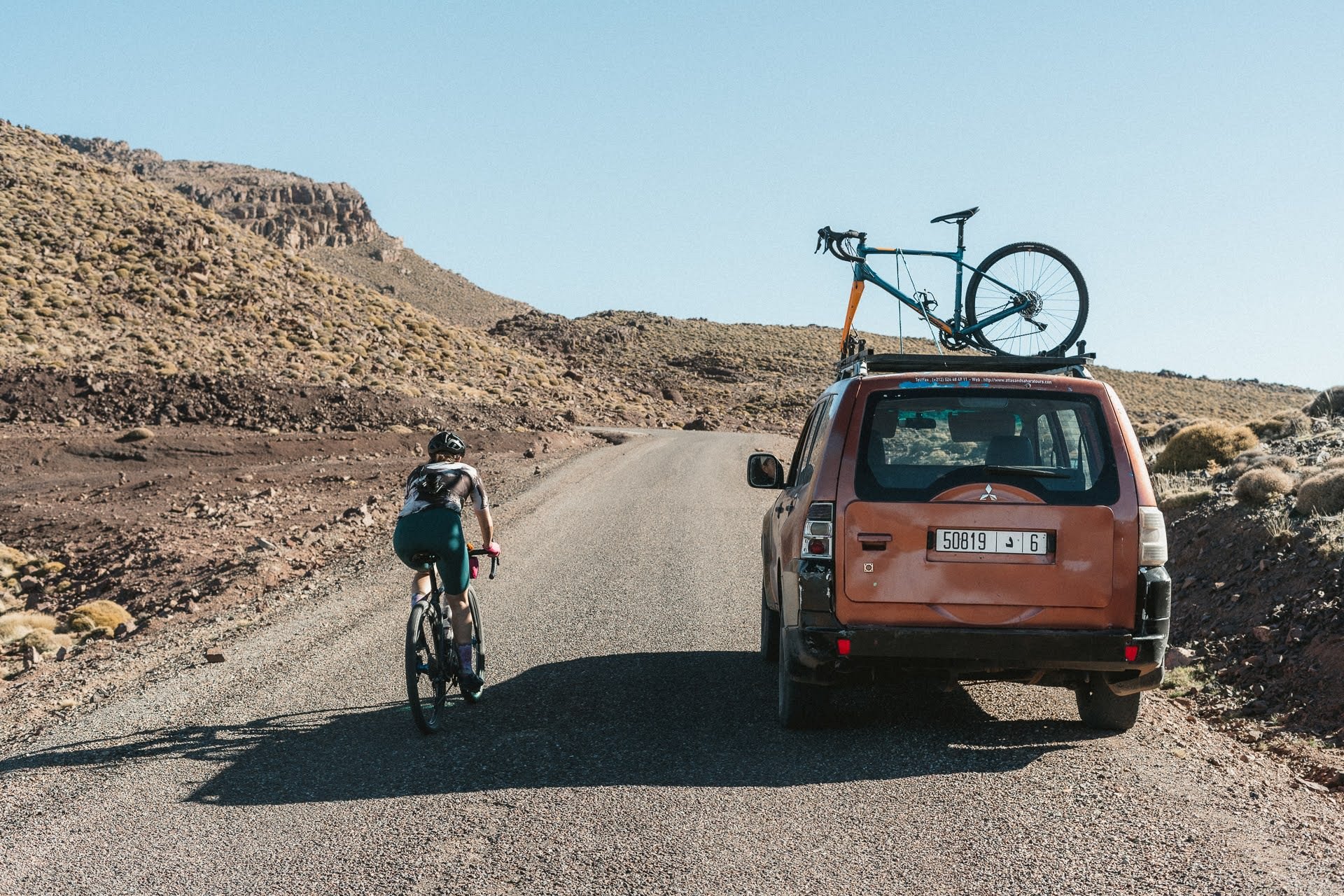 Cyclist riding on a gravel road next to an orange SUV with a bike mounted on its roof in a rocky, desert landscape.