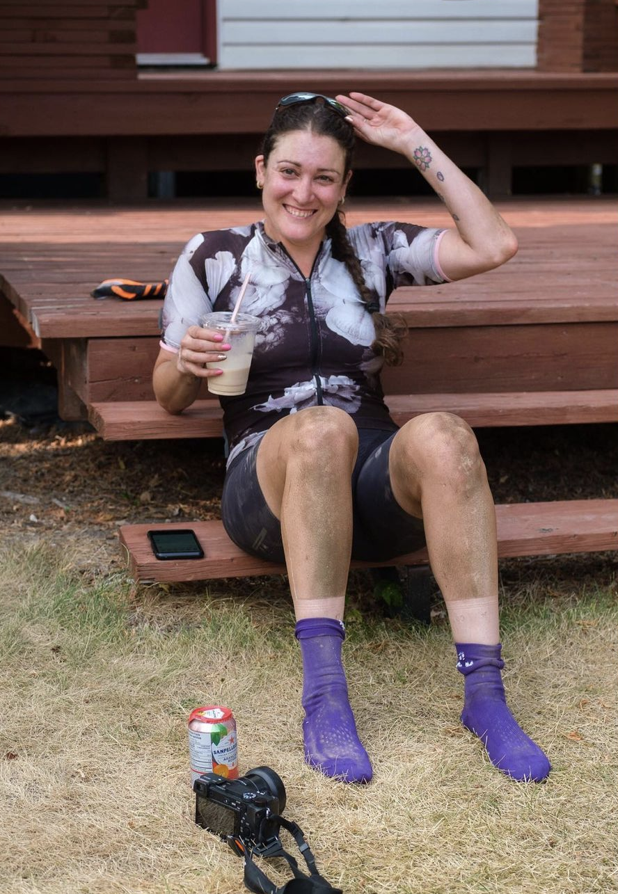 Smiling woman in a cycling outfit sitting on wooden steps, holding a drink, with purple socks and a camera on the grass nearby.