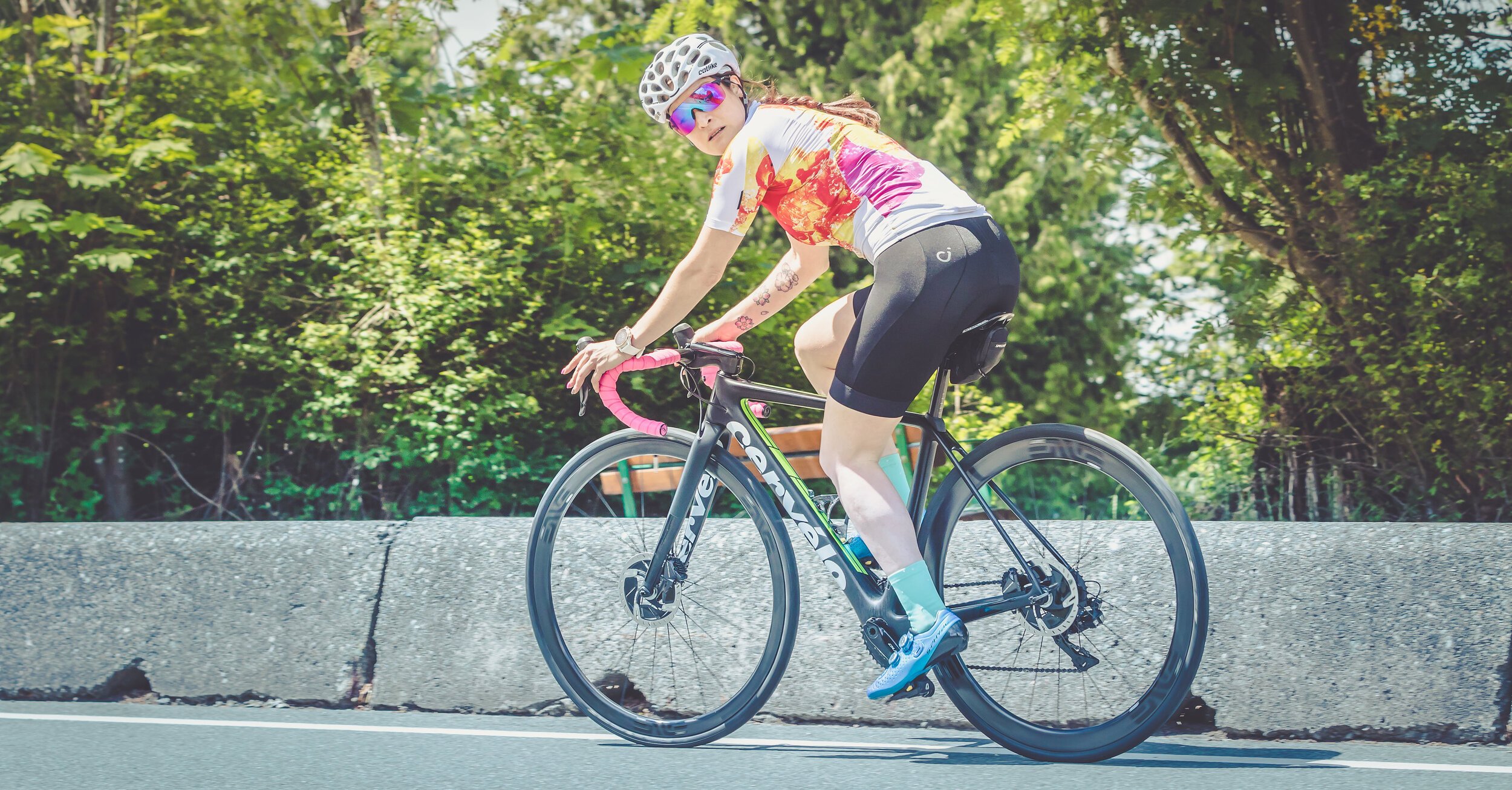 Female cyclist wearing a colorful jersey and helmet riding a black Cervelo road bike on a paved road beside a concrete barrier with green trees in the background.