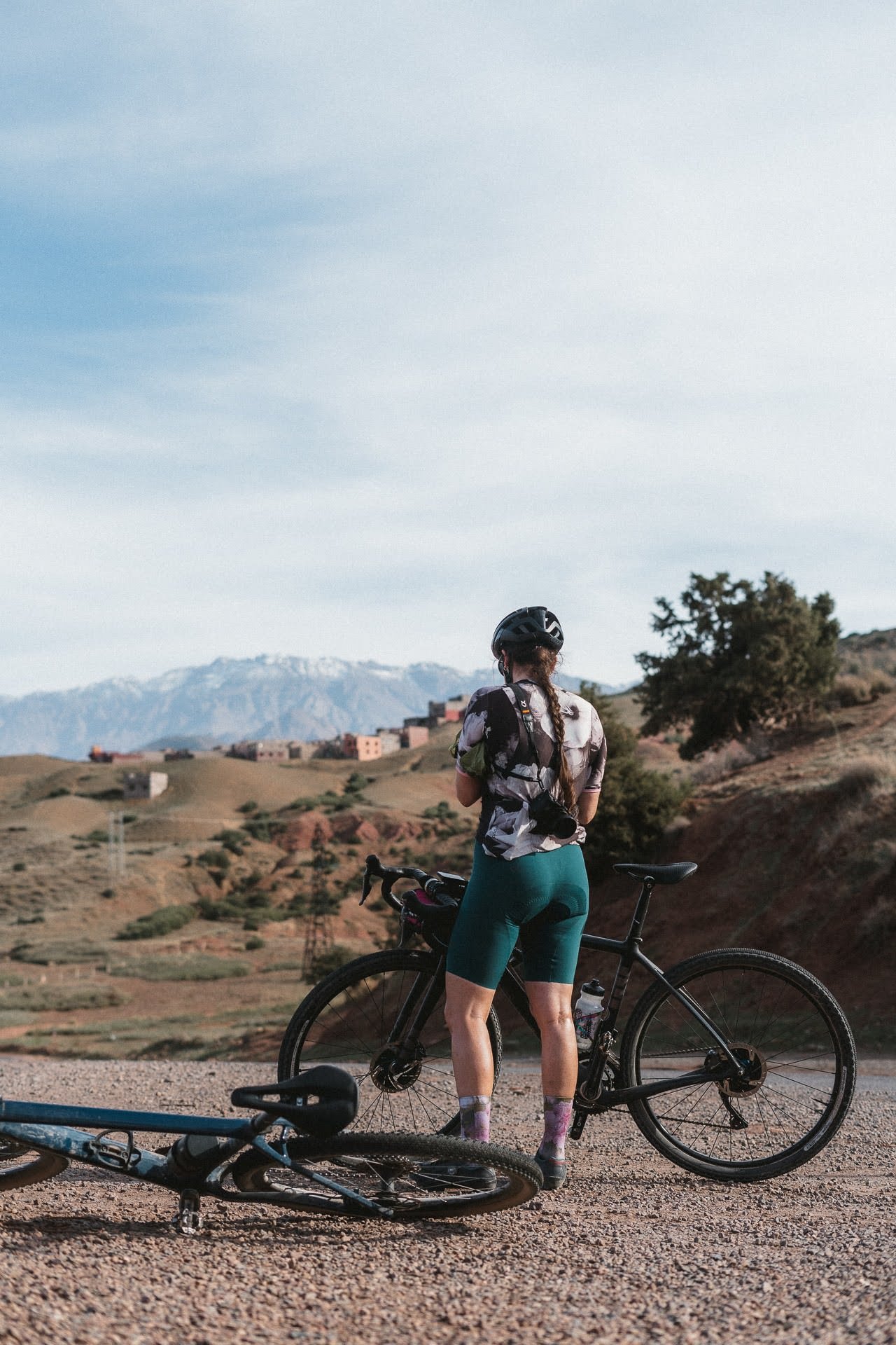 Cyclist with a helmet standing on a gravel path next to a black bike and another bike lying on the ground with mountains in the background.