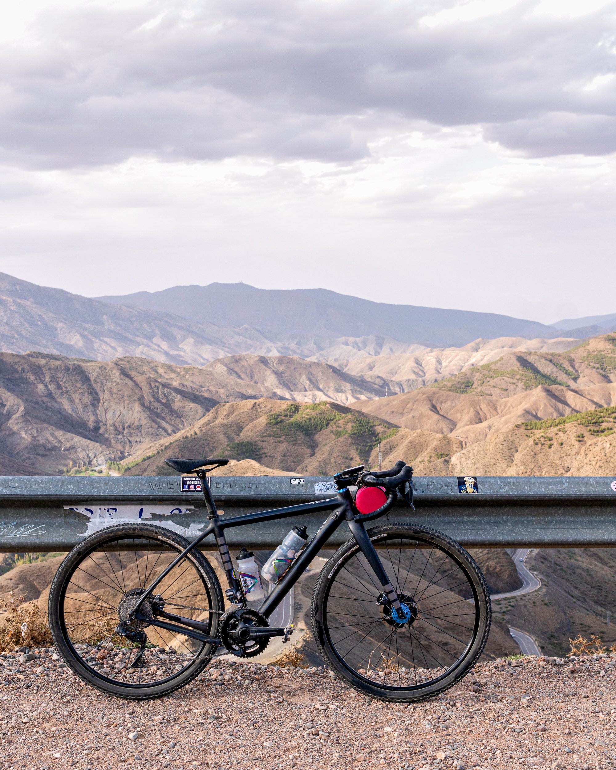 Black road bicycle parked on a gravel roadside with mountainous landscape and cloudy sky in the background.