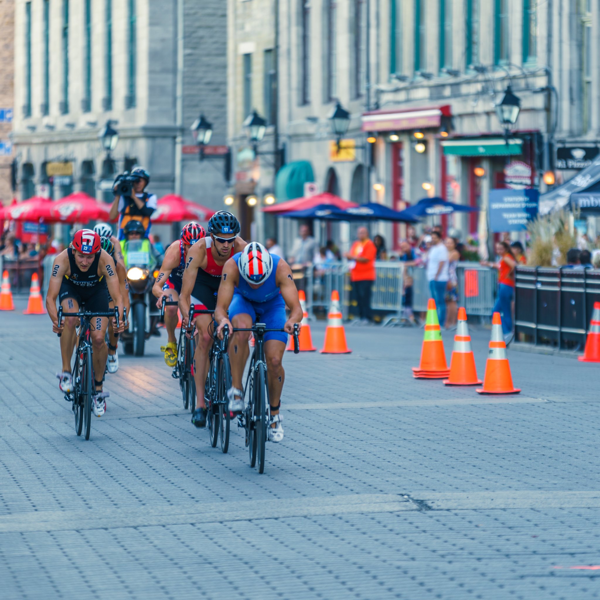 Four athletes in helmets racing on road bikes during a city cycling competition with spectators and traffic cones in the background.