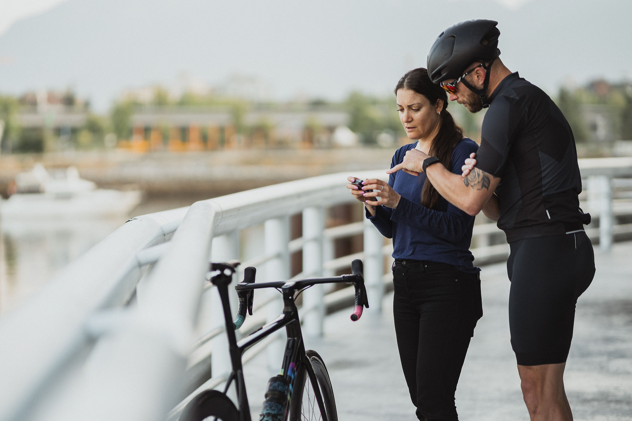 Cyclist in black gear and helmet reviewing a device with a woman on a waterfront path next to a bike.